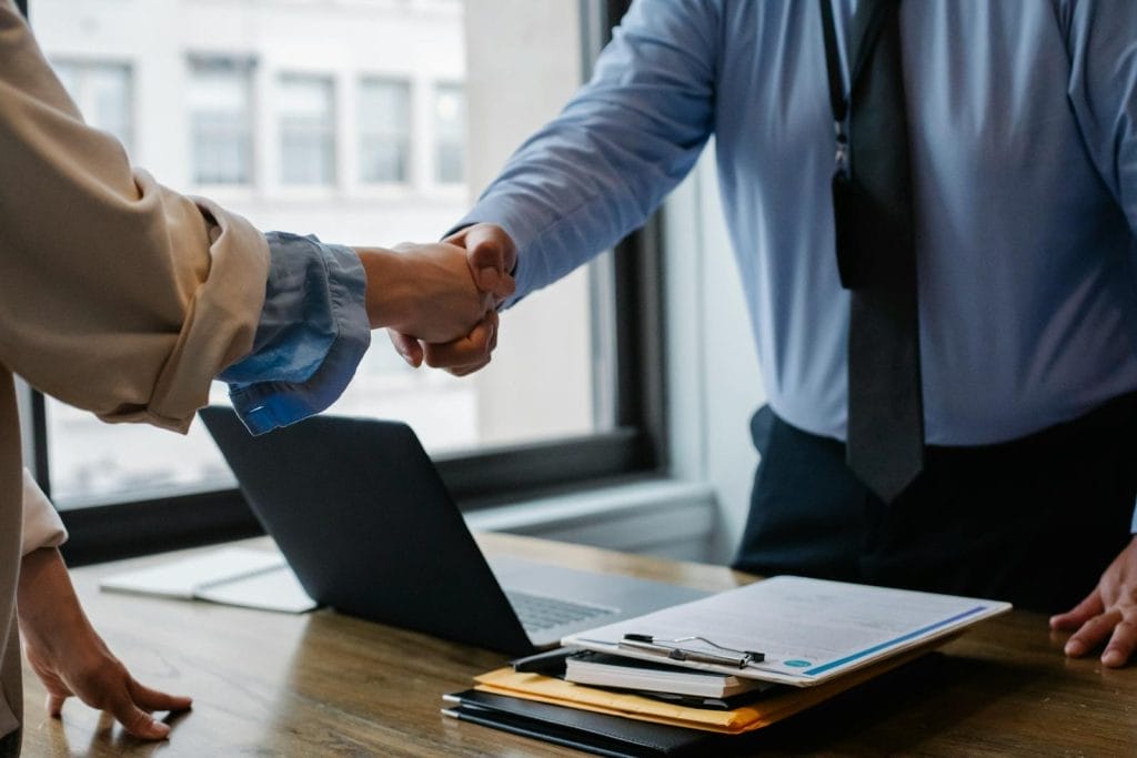 Modelo de Contrato de Patrocínio de Atleta 2 | Juristas Crop unrecognizable coworkers in formal wear standing at table with laptop and documents while greeting each other before meeting