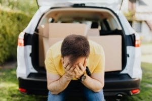 Faceless frustrated tired male wearing casual outfit sitting on car luggage boot and covering face with hands during relocation on sunny summer day